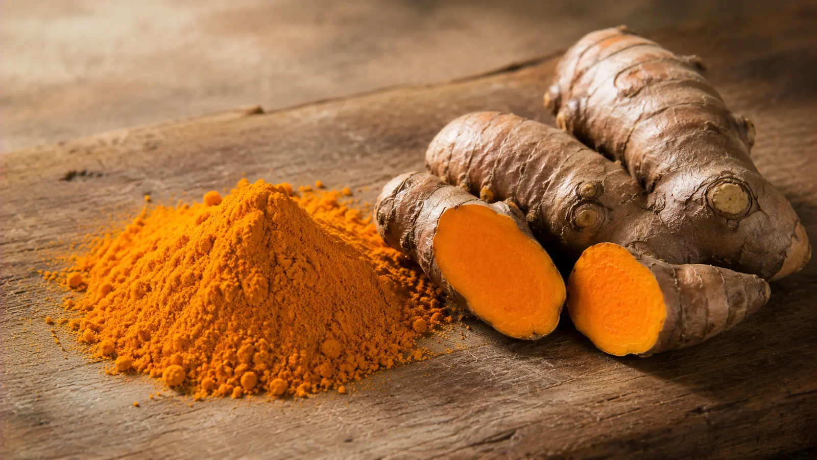 Ein Häufchen leuchtend gelb-oranges Kurkumapulver neben einer frischen Kurkumawurzel auf einem Holzbrett