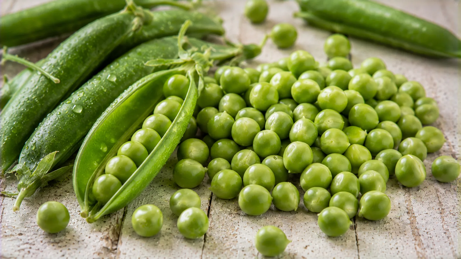 Frisch gepalte grüne Erbsen neben geöffneten Erbsenschoten auf einem hellen Holztisch